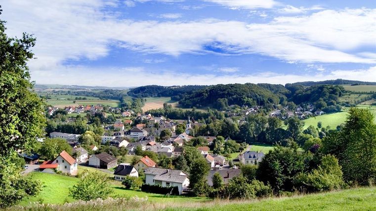Eine malerische Landschaft mit einem kleinen Dorf zwischen sanften Hügeln. Der Himmel ist klar und die Natur ist grün und einladend.