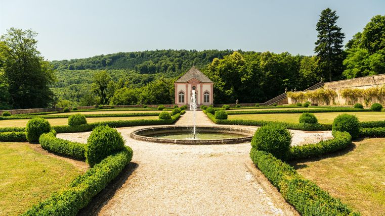 Ein schöner Garten mit geformten Buchsbaumhecken und einem zentralen Brunnen. Im Hintergrund steht ein historisches Gebäude, umgeben von Bäumen und Hügeln.