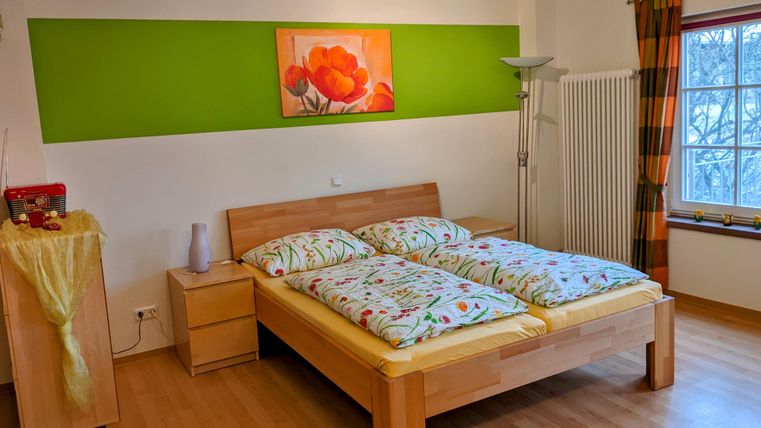 Ein helles Schlafzimmer mit einem Doppelbett und bunten Bettdecken. An der Wand befindet sich ein grüner Streifen und ein farbenfrohes Bild von Blumen.