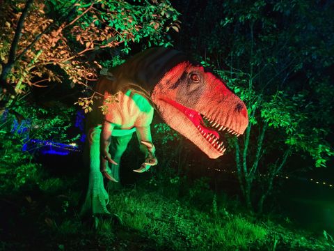 A lifelike dinosaur standing among trees, illuminated by green and red light. The scene conveys a mystical and exciting atmosphere.
