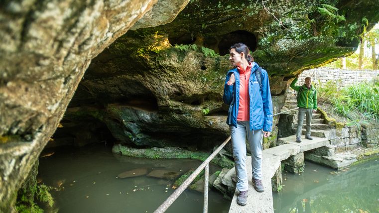 Zwei Personen stehen auf einem schmalen Steg über einem Teich, umgeben von Felsen und Vegetation.