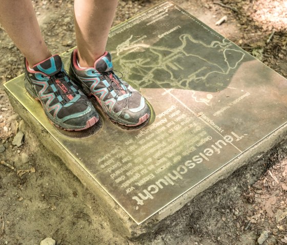 Eine Person mit bunten Sportschuhen steht auf einer Infotafel im Wald. Die Tafel zeigt eine Karte und den Text 'Teufelsschlucht'.