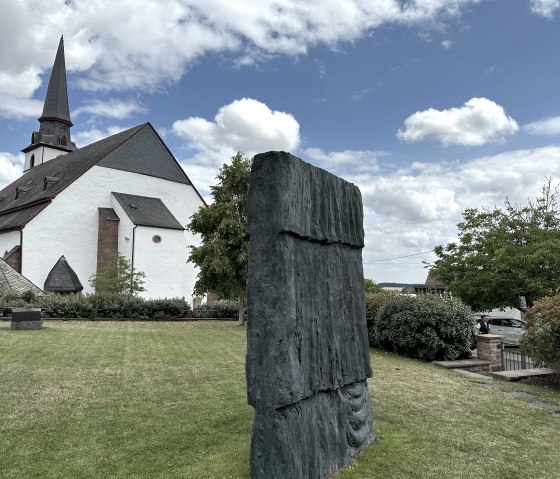 Een kerk met een spitse toren naast een modern beeldhouwwerk in de tuin van de Günther Förg bibliotheek in Weidingen., © Felsenland Südeifel, Anna Carina Krebs