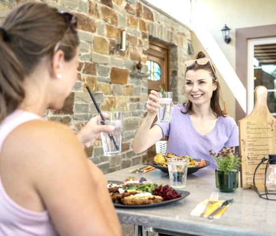 Two women sit at an outdoor table, toasting with drinks and enjoying a meal. A stone wall in the background., © Eifel Tourismus GmbH, Dominik Ketz