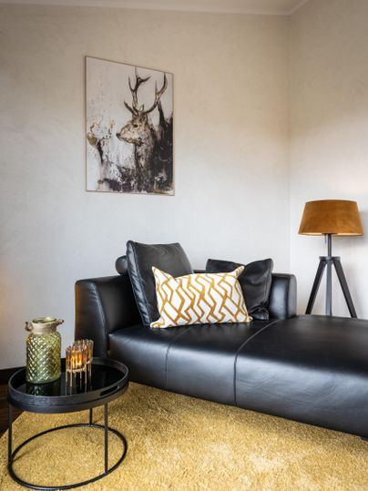 Ein modernes Wohnzimmer mit einer schwarzen Couch und einem bunten Kissen. An der Wand hängt ein Bild mit einem Hirsch und es gibt einen Stehlampe sowie einen kleinen Tisch.
