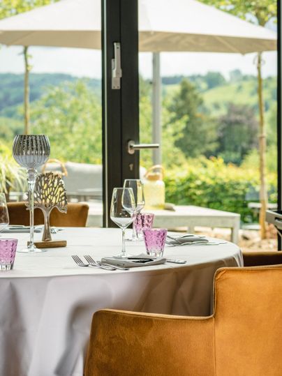 Ein eleganter Tisch mit Gläsern und dekorativen Elementen steht in einem Restaurant. Durch die Fenster sieht man eine grüne Landschaft und Sonnenschirme im Freien.