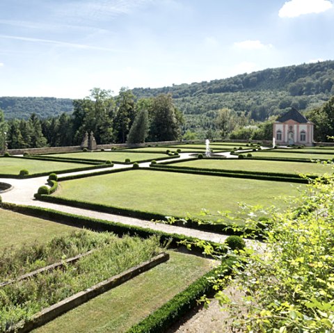 Jardin à la française avec des haies et des pelouses symétriques, au fond un petit bâtiment et des collines boisées sous un ciel bleu., © V. Teuschler