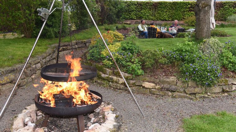 Ein gemütlicher Garten mit einem offenen Feuer in einem Grill. Im Hintergrund sitzen Menschen entspannt auf einer Wiese.