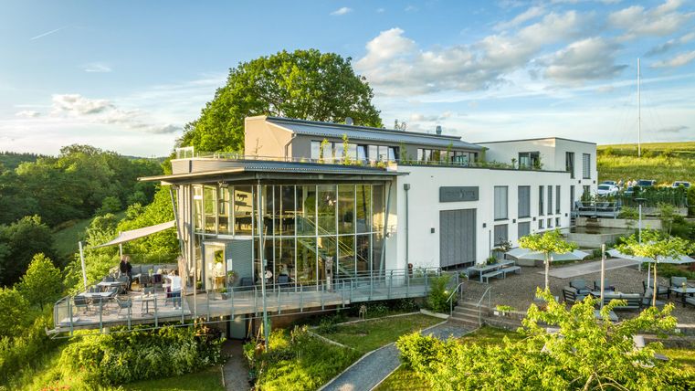 Ein modernes Gebäude mit großen Fenstern steht in einer grünen Umgebung. Die Terrasse ist ideal für entspannte Stunden im Freien.