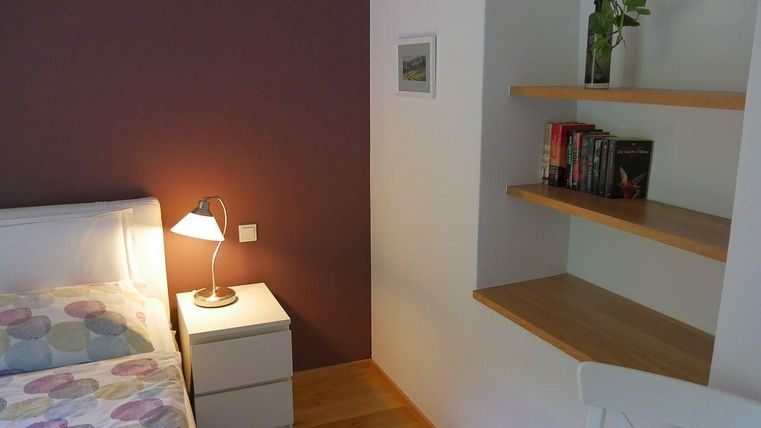 Ein gemütliches Schlafzimmer mit einem Bett, Neben Tisch und einer Lampe. An der Wand befindet sich eine helle Regalfläche mit Büchern und einer Pflanze.