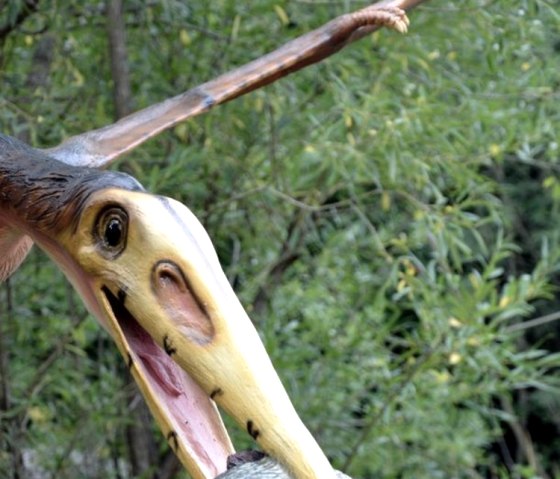 Ein Flugsaurier-Modell h&auml;lt einen Fisch im Schnabel, umgeben von gr&uuml;ner Vegetation im Dinopark.