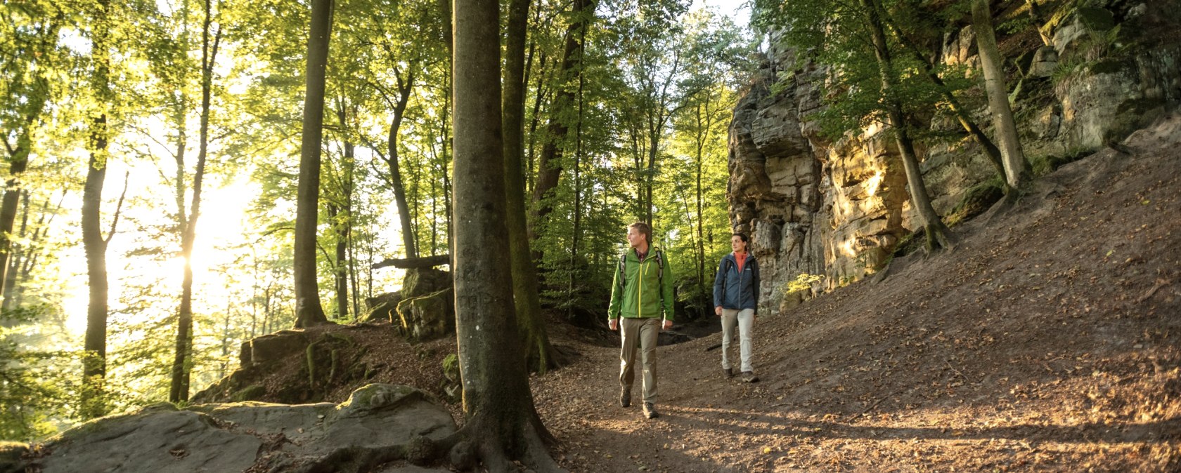 Two hikers are walking on a forest path along a rock face. The sun shines through the trees and bathes the scene in warm light., © Dominik Ketz