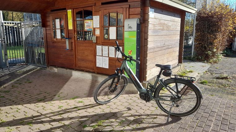 Ein Elektrofahrrad steht vor einem Holzgebäude mit Fenstern und einer Informationstafel. Das Wetter ist sonnig und der Boden ist mit etwas Laub bedeckt.