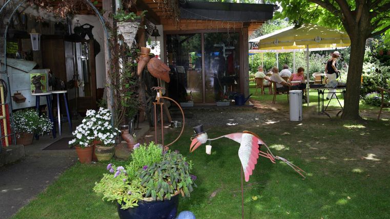 Ein einladender Garten mit bunten Pflanzen und einem fließenden Brunnen. Im Hintergrund sind Sitzplätze unter einem Sonnenschirm und eine gemütliche Atmosphäre zu sehen.