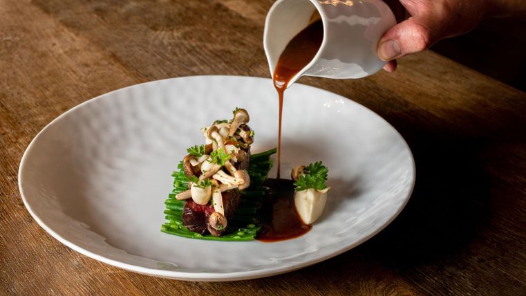 Ein kunstvoll angerichteter Teller mit Gemüse und Sauce. Eine Hand gießt die Sauce über die Speise.