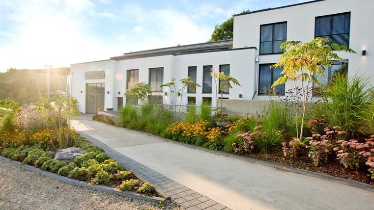 Ein modernes Gebäude mit großen Fenstern und einem einladenden Eingang. Der Garten ist üppig mit bunten Blumen und grünen Pflanzen gestaltet.