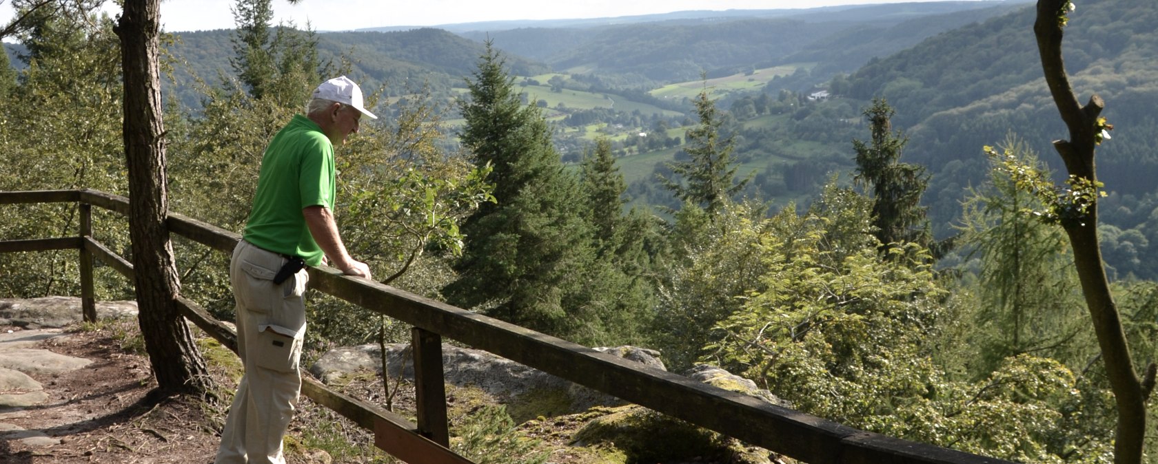 A man in green clothing stands at a railing and looks out over a wooded landscape with hills and valleys., &copy; Lauschtour.de