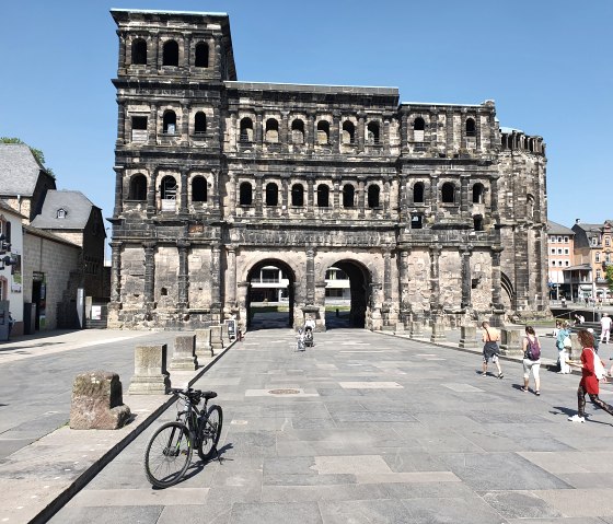 Die Porta Nigra in Trier bei sonnigem Wetter. Menschen spazieren, ein Fahrrad steht im Vordergrund. Historische Architektur vor blauem Himmel., © TI Bitburger Land, Steffi Wagner