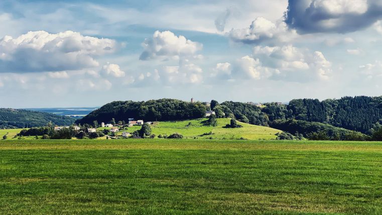 Eine malerische Landschaft mit grünen Wiesen und sanften Hügeln. Der Himmel ist blau mit einigen weißen Wolken.