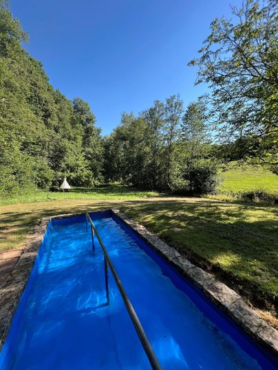 Ein blauer Pool steht in einer grünen Wiese, umgeben von Bäumen und Sträuchern. Im Hintergrund sind einige Zelte sichtbar.