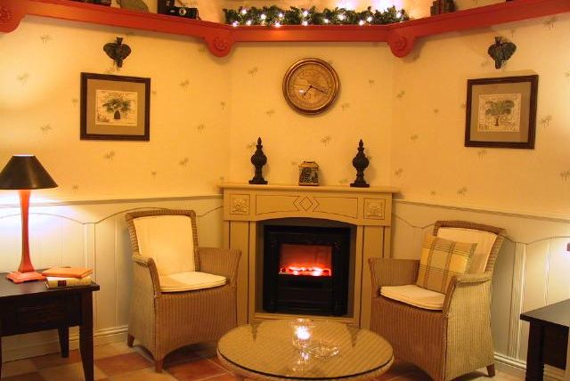 Ein gemütlicher Raum mit einem Kamin, zwei bequemen Stühlen und einem runden Tisch. Die Wände sind mit Bildern dekoriert und es gibt eine warmes Lichtambiente.