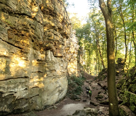 Two hikers walk through the Devil's Gorge, flanked by high rock faces and surrounded by dense forest in the sunlight., &copy; Dominik Ketz