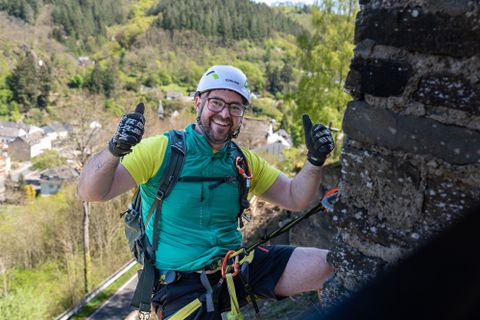 Ein Kletterer mit Helm und Sicherheitsausrüstung lächelt und zeigt Daumen nach oben. Im Hintergrund sind grüne Hügel und ein kleines Dorf zu sehen.