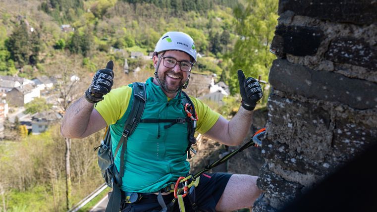 Ein Kletterer mit Helm und Sicherheitsausrüstung lächelt und zeigt Daumen nach oben. Im Hintergrund sind grüne Hügel und ein kleines Dorf zu sehen.