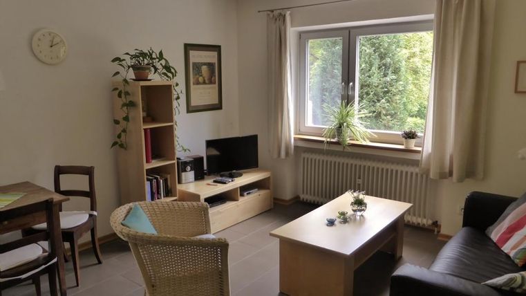 Ein gemütliches Wohnzimmer mit einem Tisch, Stühlen und einem schwarzen Sofa. Große Fenster lassen viel Licht herein und bieten einen Blick ins Grüne.