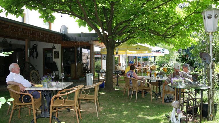 Ein gemütliches Café im Freien mit mehreren Tischen und Stühlen unter einem großen Baum. Gäste genießen die entspannte Atmosphäre in einem grünen Garten.
