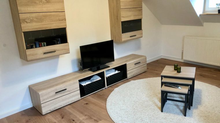 Ein modernes Wohnzimmer mit einem Fernseher und Holzregalen an der Wand. Der Boden ist aus Holz, und ein runder Teppich sowie ein kleiner Tisch sind vorhanden.