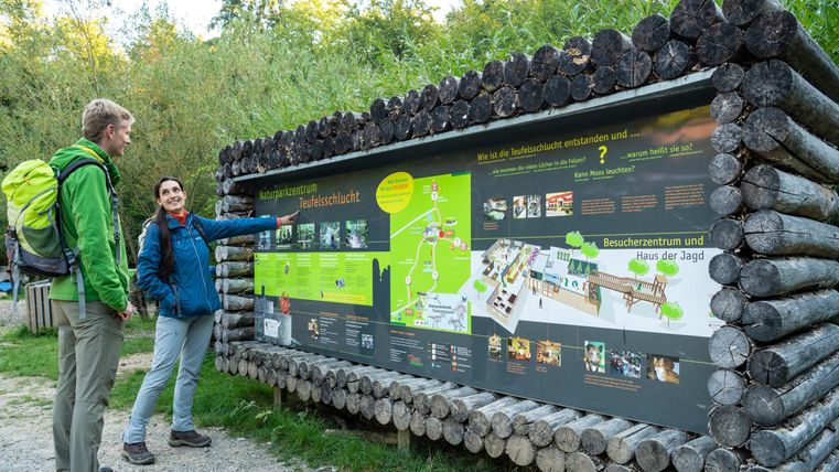 Ein Paar steht vor einem Informationsschild im Wald. Das Schild zeigt eine Karte und Informationen über die Umgebung und Aktivitäten.