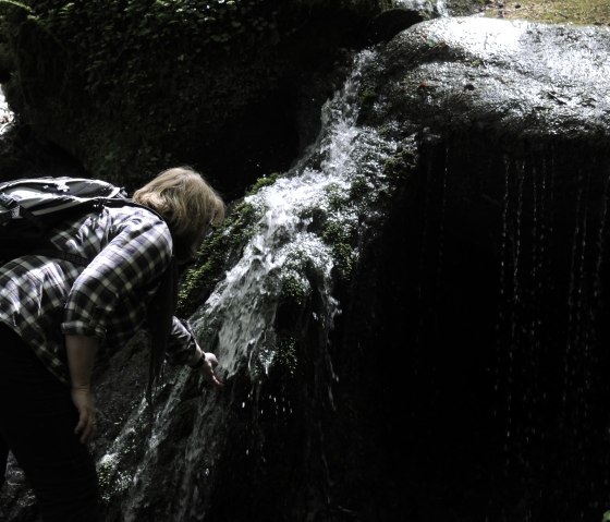 Eine Person mit Rucksack beugt sich über einen kleinen, moosbedeckten Wasserfall im Wald., © Felsenland Südeifel Tourismus GmbH