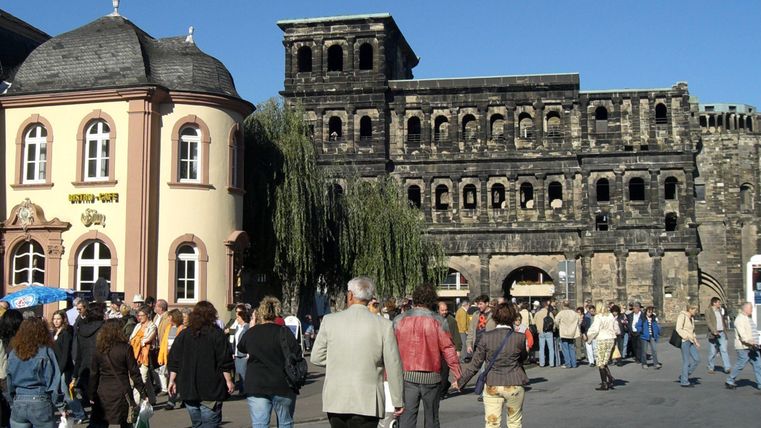 Ein belebter Platz mit vielen Menschen und historischen Gebäuden. Im Hintergrund steht eine alte Steinfassade.