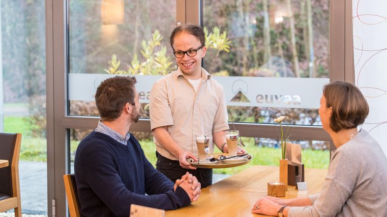 Ein Kellner bringt Getränke zu einem Tisch mit zwei Gästen in einem modernen Café. Die Atmosphäre ist freundlich und einladend.