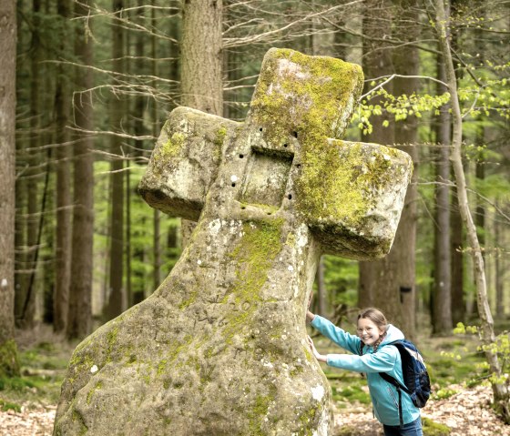 Mädchen in blauer Jacke lehnt sich an das moosbedeckte Fraubillenkreuz im Wald von Bollendorf., © Eifel Tourismus GmbH, Dominik Ketz
