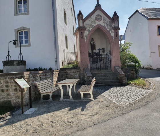 Kleine Kapelle mit Statue und Sitzbank in Wolsfeld. Ein Brunnen und Informationstafel sind ebenfalls zu sehen. Sonniger Tag., © Tourist-Information Bitburger Land, Steffi Wagner