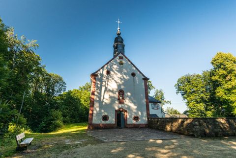 Eine kleine Kirche mit einem Turm und einem Kreuz an der Spitze. Umgeben von grünen Bäumen und einem klaren blauen Himmel.