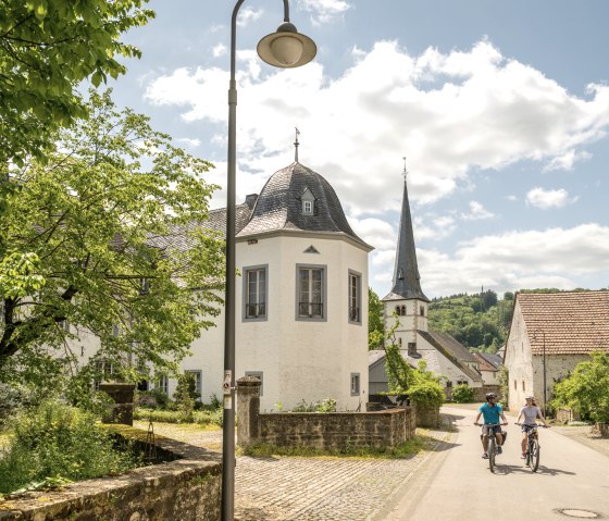 Deux cyclistes roulent sur une route ensoleillée à Wolsfeld. A gauche, des bâtiments historiques et un arbre. A l'arrière-plan, on aperçoit une église., © Eifel Tourismus GmbH, Dominik Ketz