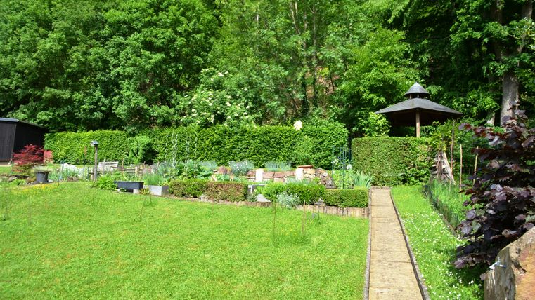 Ein schöner Garten mit grünem Gras und dichtem Gestrüpp. Im Hintergrund sieht man ein Pavillon und beete mit verschiedenen Pflanzen.