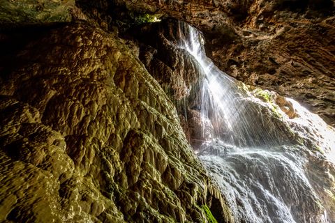 Ein Wasserfall fließt über eine steinige Felswand in einer dunklen Höhle. Das Licht reflektiert sich auf dem Wasser und sorgt für eine geheimnisvolle Atmosphäre.