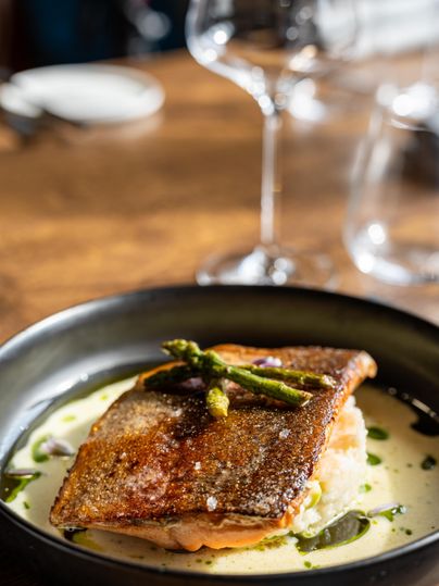 Ein köstliches Gericht mit knusprigem Fischfilet auf einer cremigen Sauce, garniert mit Spargel. Im Hintergrund sind eine weinartige Gläser und ein rustikaler Tisch sichtbar.