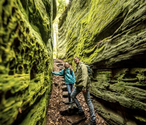 Two hikers in a narrow, moss-covered gorge. The walls are green and steep, the ground is covered in leaves. The scene looks adventurous., © Eifel Tourismus GmbH, Dominik Ketz