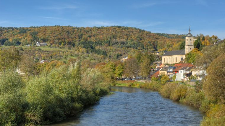 Ein ruhiger Fluss fließt durch eine malerische Landschaft mit bunten Bäumen im Hintergrund. Im Bild ist eine kleine Stadt mit einer Kirche zu sehen.