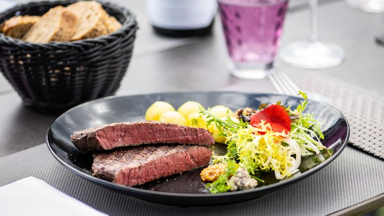 Ein schöner Teller mit saftigem Steak, Beilagen aus Kartoffeln und frischem Salat. Im Hintergrund sind Brot und ein Getränk zu sehen.