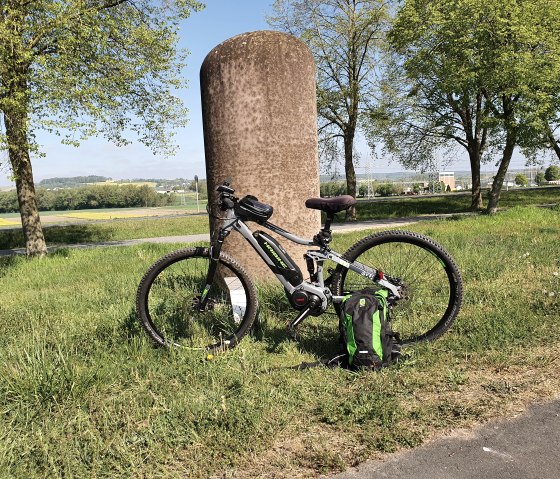 Ein Fahrrad lehnt an einer Steinsäule auf einer grünen Wiese. Im Hintergrund sind Bäume und eine weite Landschaft zu sehen., © TI Bitburger Land, Steffi Wagner