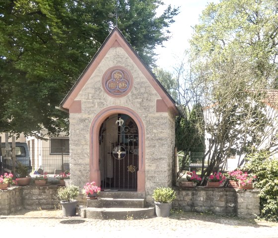Eine kleine Steinkapelle mit spitzem Dach, umgeben von Blumen und Bäumen. Ein Straßenschild zeigt 'Meilbrücker Straße' an., © TI Bitburger Land