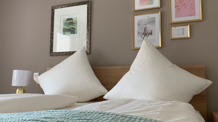 Ein gemütliches Schlafzimmer mit einem Holzbett und großen Kopfkissen. An der Wand hängen dekorative Bilder in Rahmen und ein Nachttisch mit einer Lampe ist sichtbar.