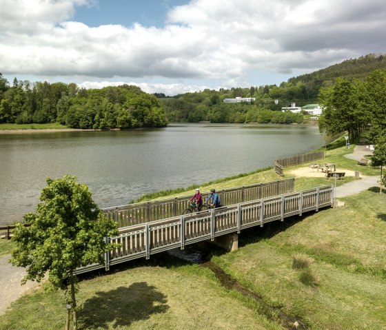 Two cyclists cross a wooden bridge at the Bitburg reservoir, surrounded by green countryside and a cloudy sky., &copy; Eifel Tourismus GmbH, Dominik Ketz