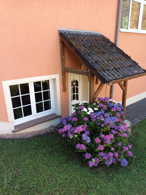 Entrée de l'appartement de vacances avec une terrasse et un auvent. Des hortensias colorés fleurissent devant une fenêtre.